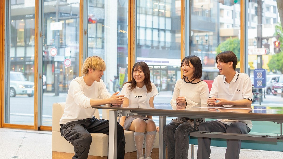 大きな窓の前のテーブルに座り、談笑している四人の若者たち。明るい自然光が差し込むカフェのような室内（画像をクリックするとページへ移動します）。