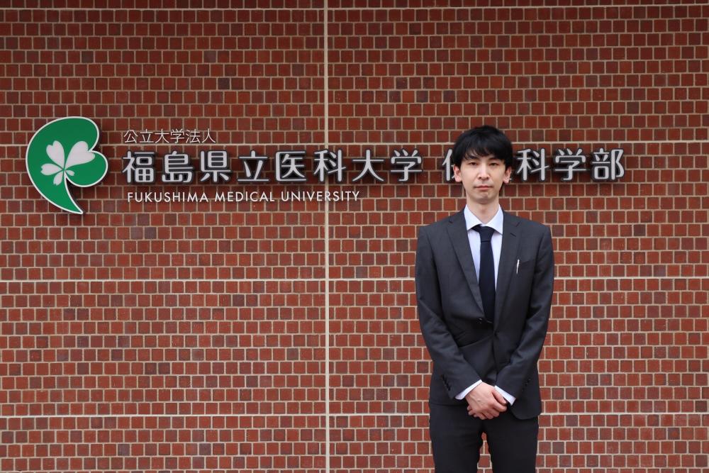福島県立医科大学の看板の前に立つ松本大典さんの写真。